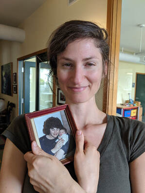 A white woman in a dark forest green t-shirt is smiling and holding a framed photo of an adult holding her as an infant.