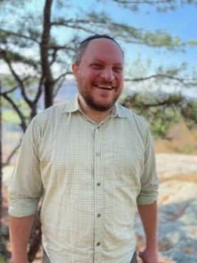 A white man wearing a black kippah is joyfully smiling. He has a short beard and is wearing a beige button up shirt. In the background there is a pine tree and bedrock with a blurred vista behind.