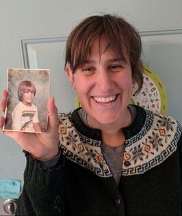 A white woman with light brown bangs wearing a black and geometric sweater is holding a paper photo of a blond child with a bowl cut wearing a rainbow t-shirt.